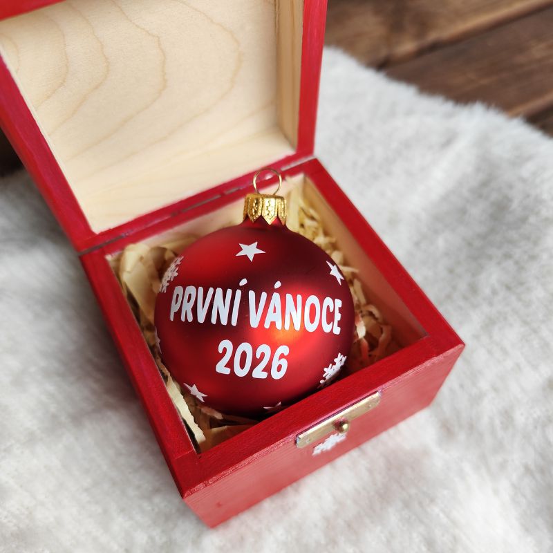 Vánoční červená baňka s nápisem První vánoce 2026 v otevřené krabičce